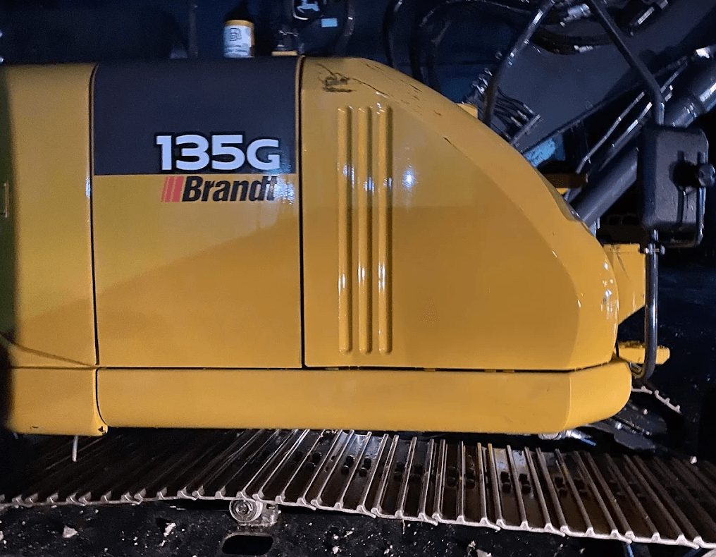 Close-up of a yellow Brandt 135G excavator with metal tracks in a dark setting.