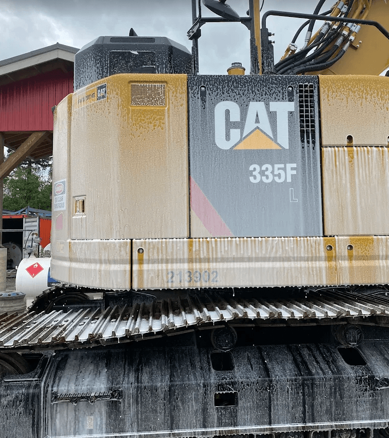 A yellow CAT 335F L excavator covered in thick white soap foam during cleaning.