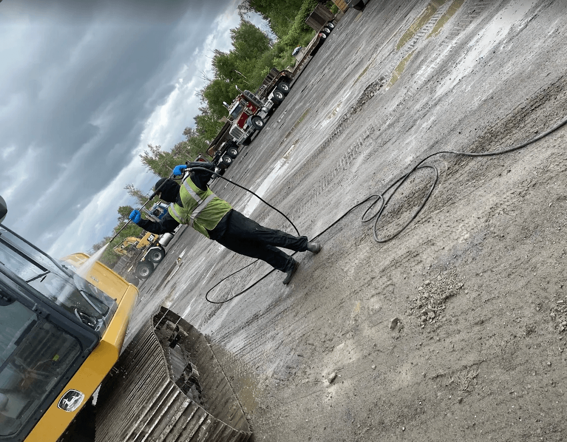 Worker in safety gear pressure washing a yellow excavator on a muddy industrial lot.